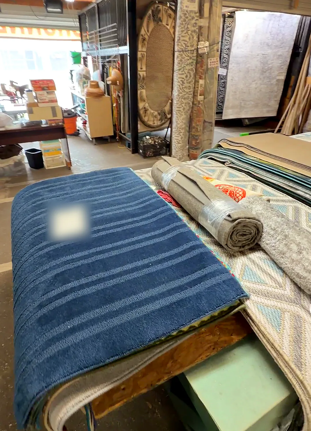 A folded navy striped rug on a table beside rolled rugs; its tag is blurred.